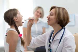 A doctor in a white coat with a stethoscope around her neck uses a pen light to look into the mouth of a young girl.
